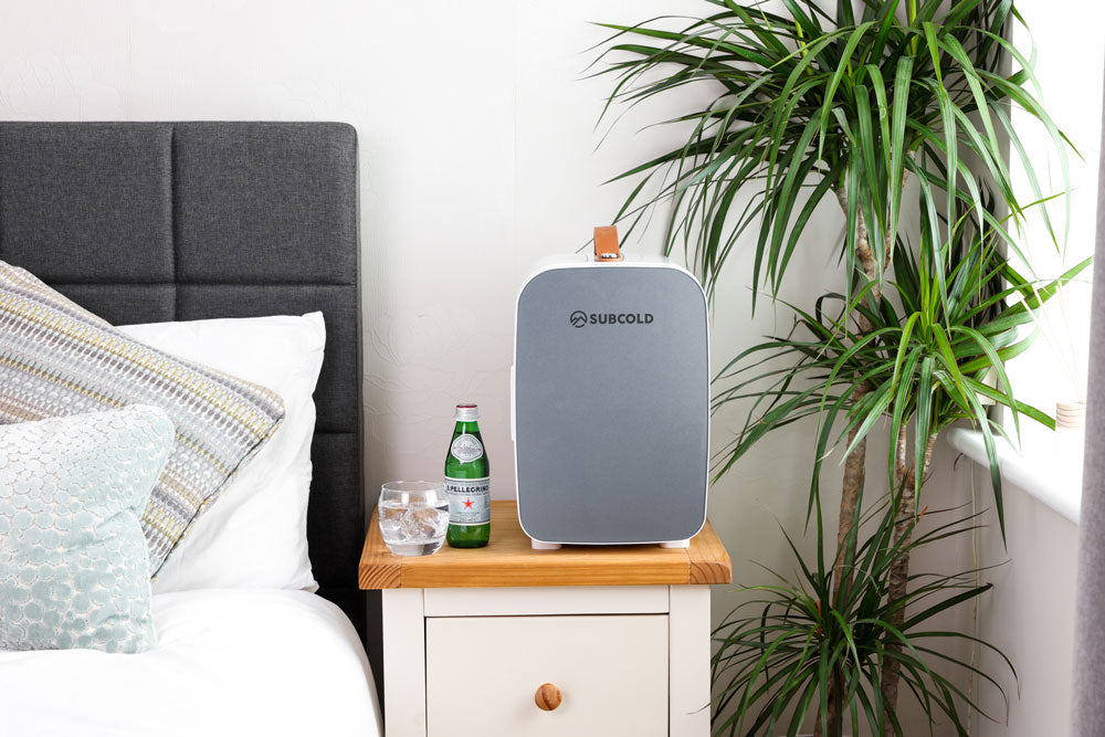 Portable mini fridge with grey front and tan handle on a bedside table