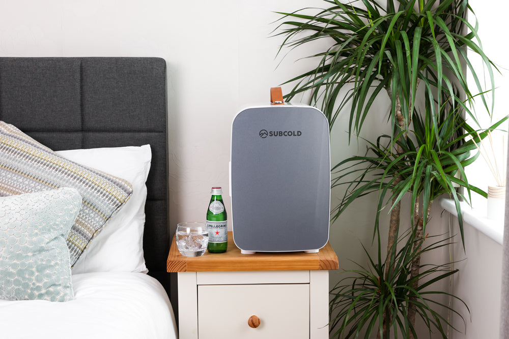 Portable mini fridge in white and dark grey on a bedside table with drinks nearby