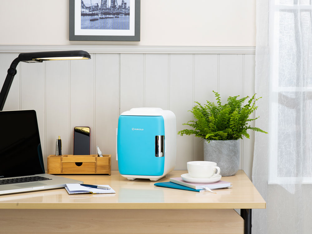 Blue portable mini fridge on a desk with cup and plant nearby