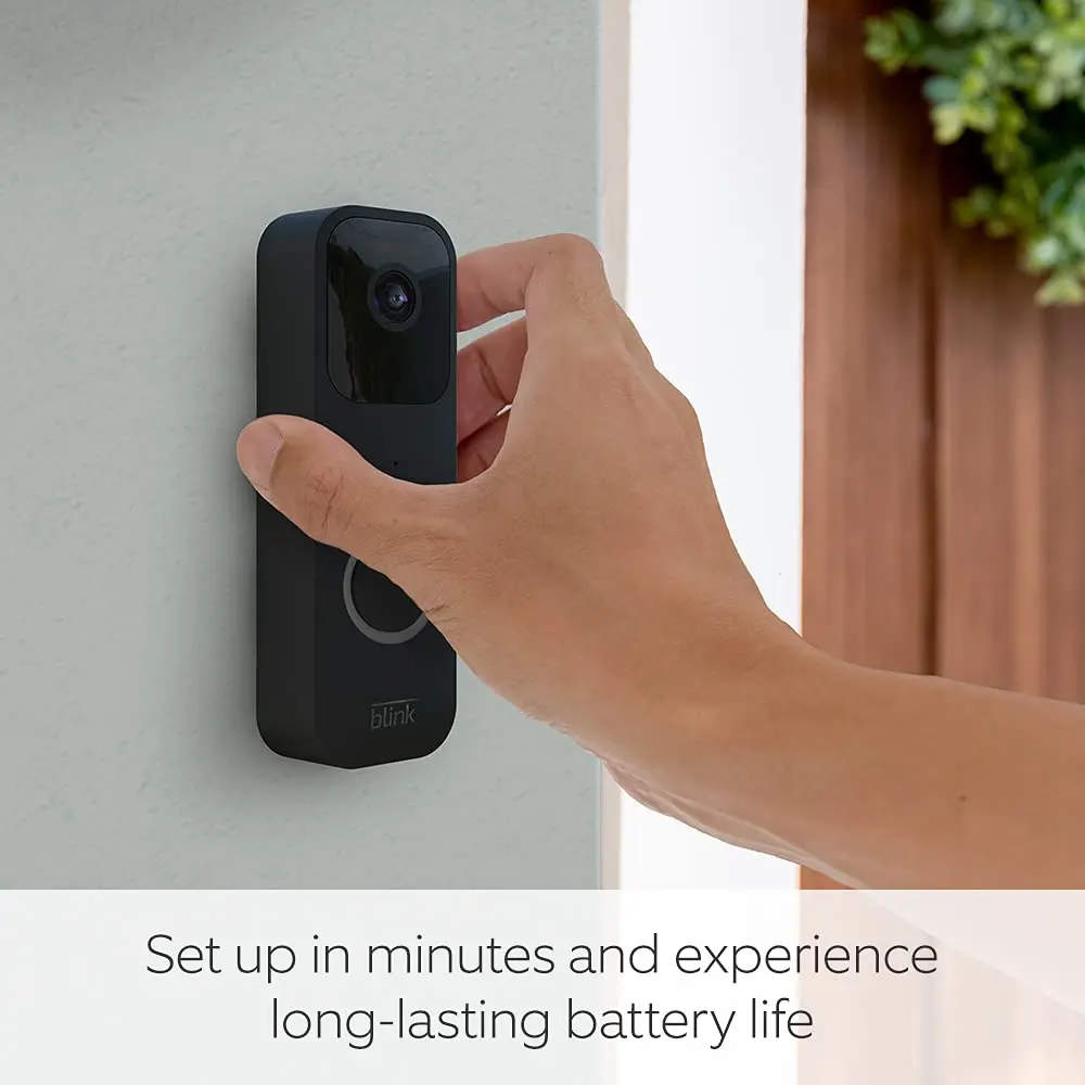 Hand holding a black video doorbell with blue ring on an outdoor wall