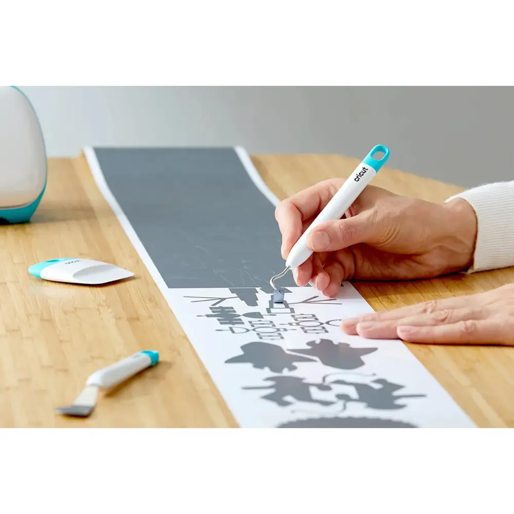 Hand using white-and-teal Cricut tool to apply design on light vinyl sheet