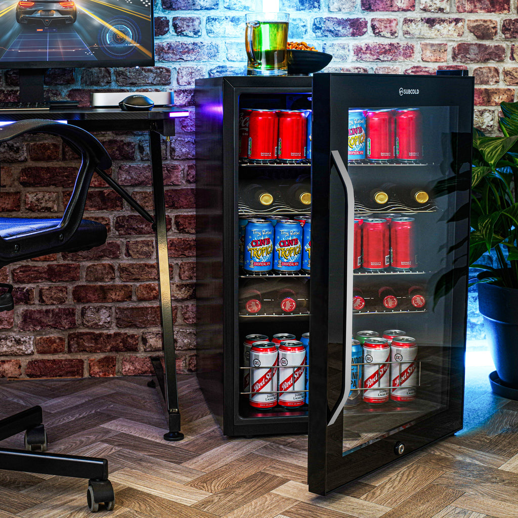 Black freestanding beer fridge with door open, LED-lit cans and bottles inside against brick wall