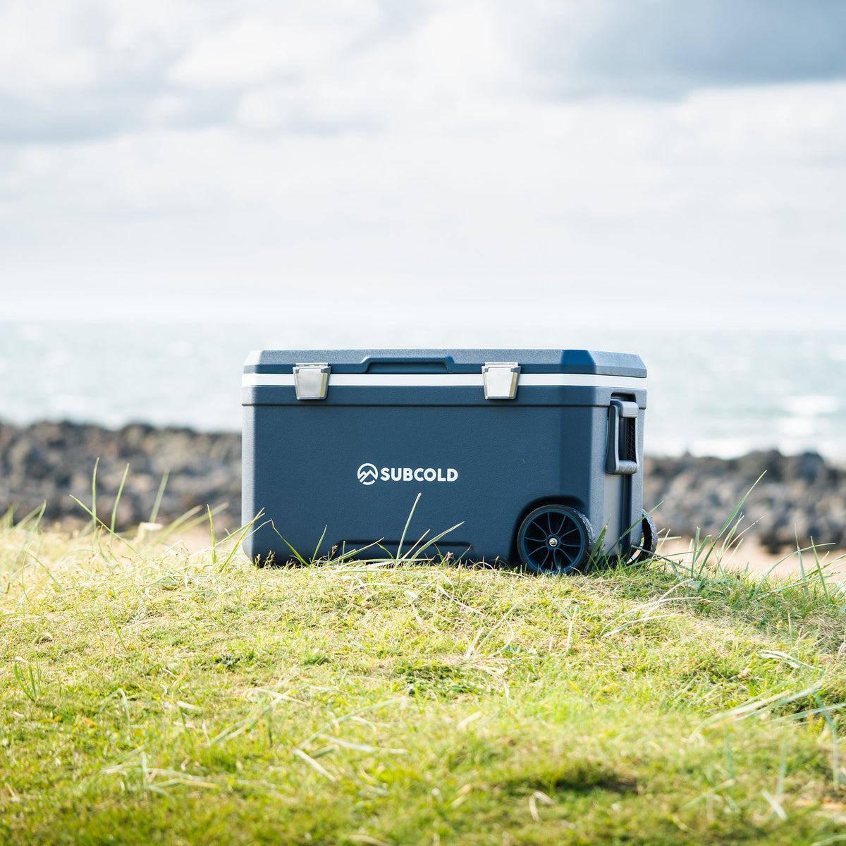 Blue wheeled cool box with closed lid and side wheel, outdoors on grass