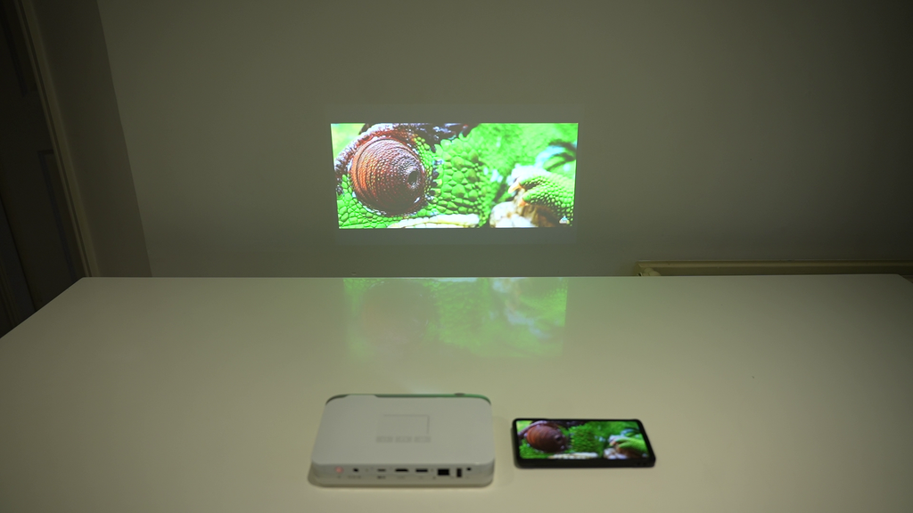 Ultra portable smart projector with phone beside it, projecting video on a screen